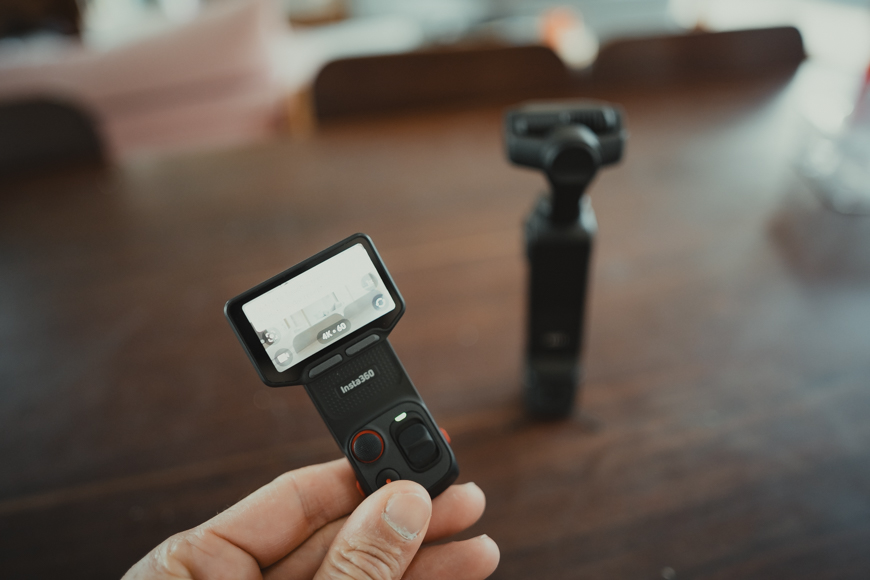 A hand holds a small handheld camera with a built-in screen; another similar device stands on a blurred wooden table in the background.