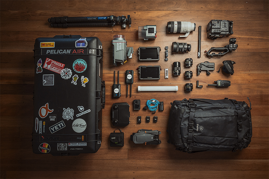 Flat lay of camera gear on a wooden floor, including a Pelican case, lenses, cameras, tripod, drone, batteries, and a black backpack.