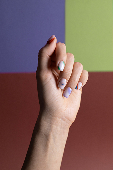 A hand with light skin tone shows off manicured nails with minimalist, multicolored designs against a geometric, colorful background.
