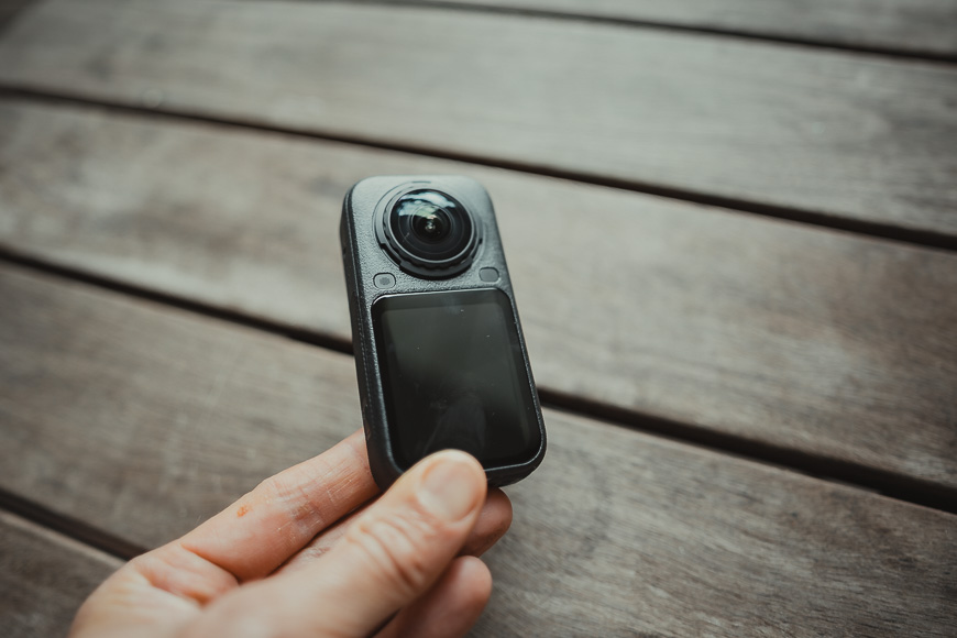 A hand holds a compact, rectangular 360-degree action camera with a prominent spherical lens.