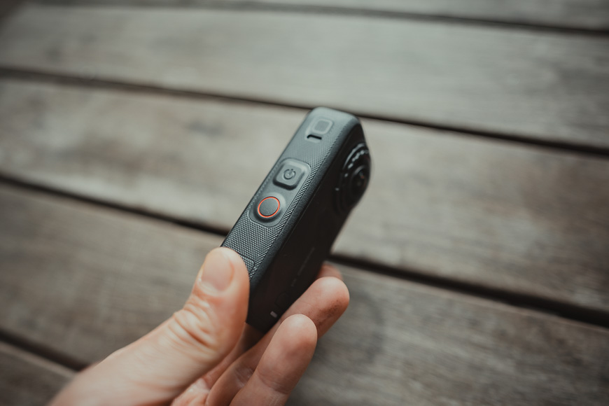 A hand holding a compact action camera with a power button and a red-rimmed shutter button visible on the side.