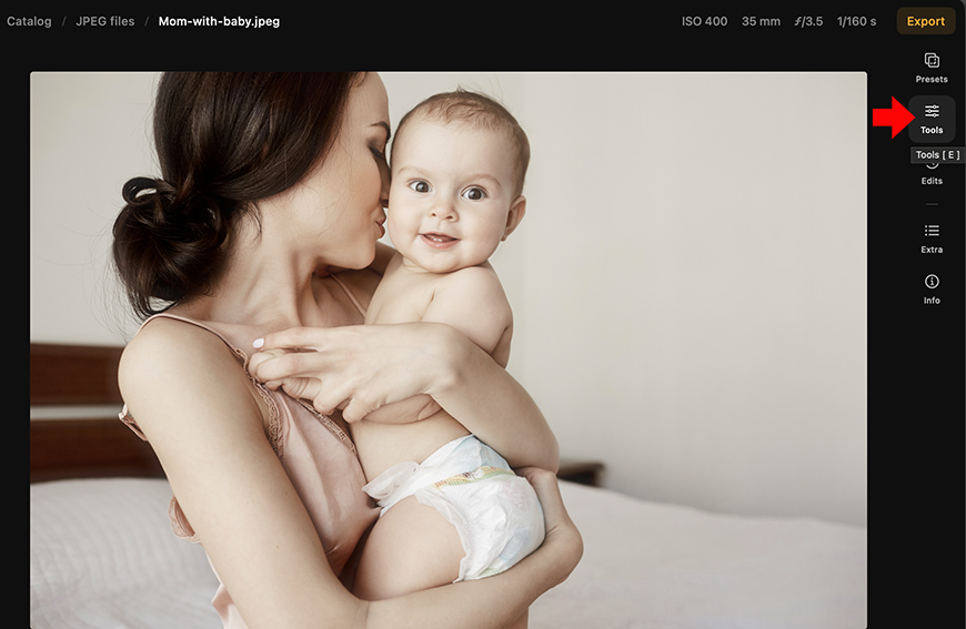 A woman holds a baby in a diaper while standing in a bedroom. Editing software interface with a red arrow pointing to a “Tools” menu is visible on the right.