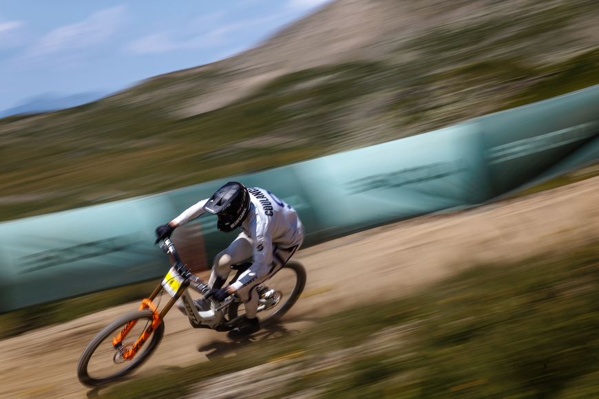 Downhill mountain biker in white jersey and black helmet rounding a dirt track corner at speed, with motion blur in background.