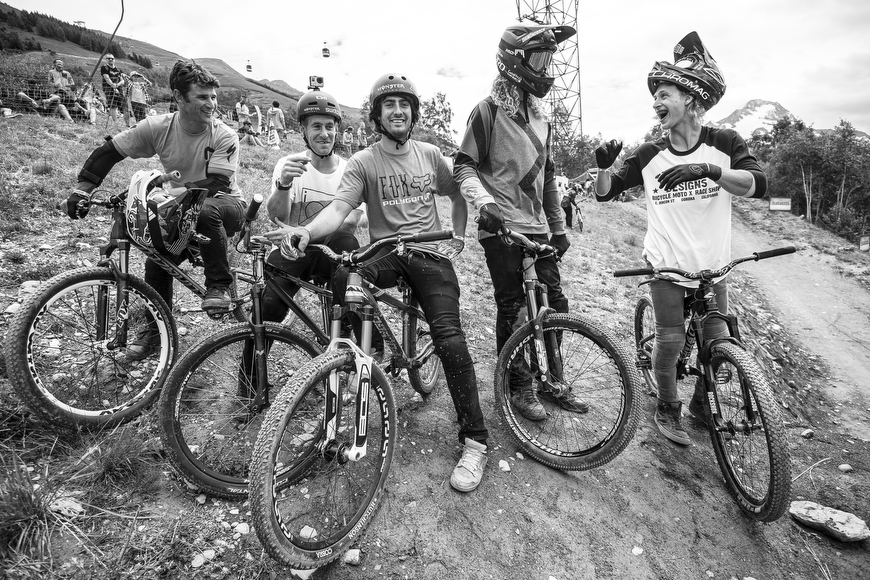 Five mountain bikers wearing helmets and gear sit on their bikes, smiling and talking on a dirt trail with trees and a hillside in the background.