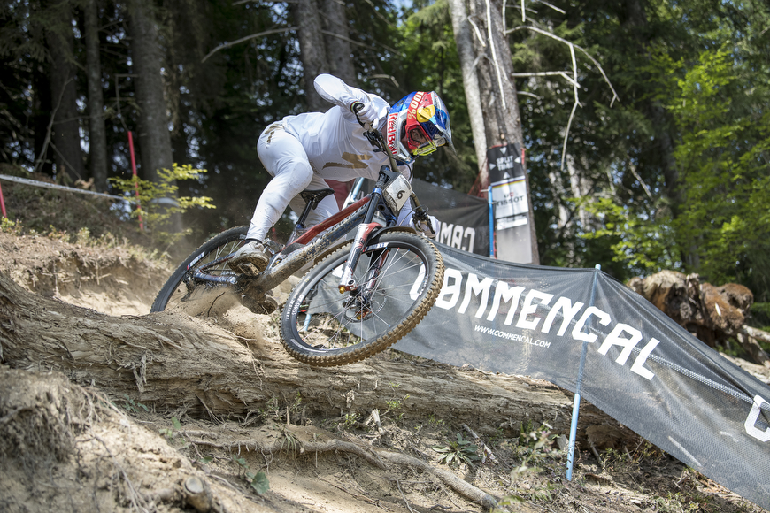 A mountain biker in white gear rides downhill over tree roots on a dirt trail, with branded barriers and forest trees in the background.