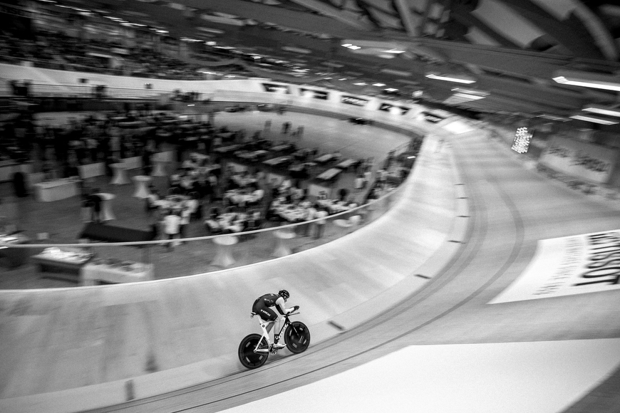 A cyclist races alone on an indoor velodrome track, with blurred spectators and tables visible in the background.