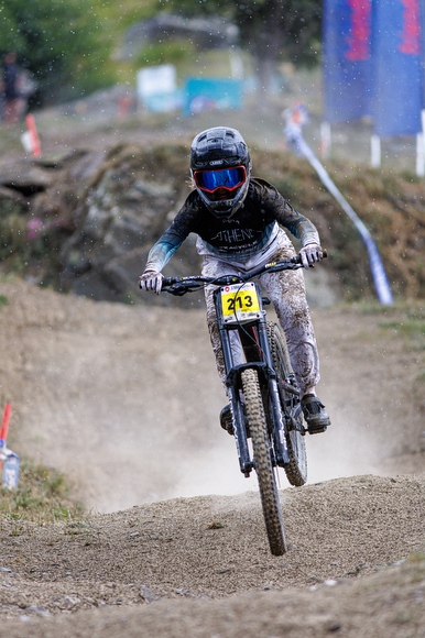 Mountain biker wearing helmet and goggles rides a dirt trail at speed, with mud splatters on clothing and a yellow race number “213” on the bike.