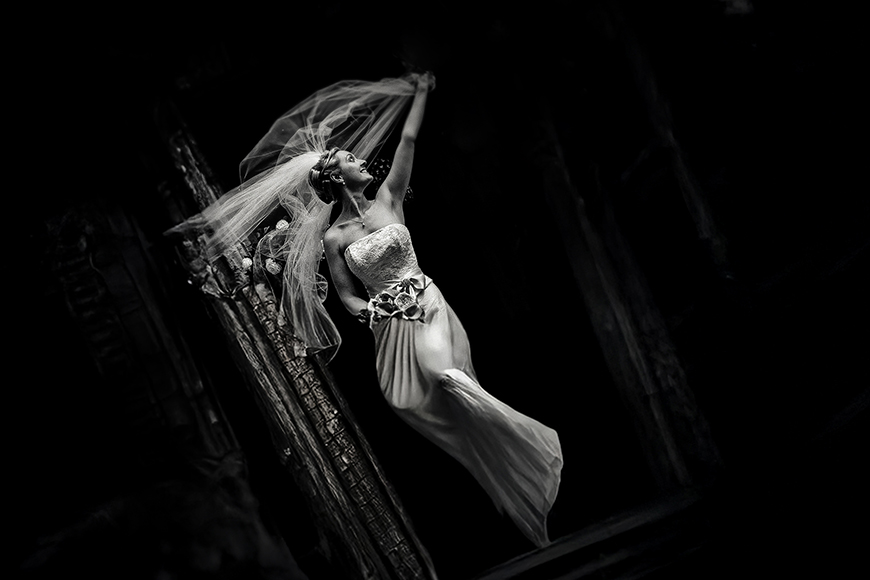 A bride in a strapless wedding gown and veil poses with one arm raised, standing at an angle against a dark background.