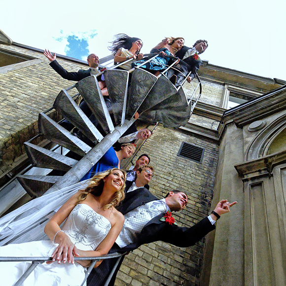 A wedding party poses on a spiral staircase outside, with the bride and groom at the bottom, and guests in formal attire standing on the steps above.