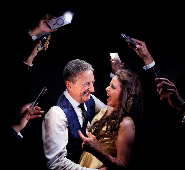 A smiling man and woman embrace while several hands around them take photos with smartphones against a dark background.