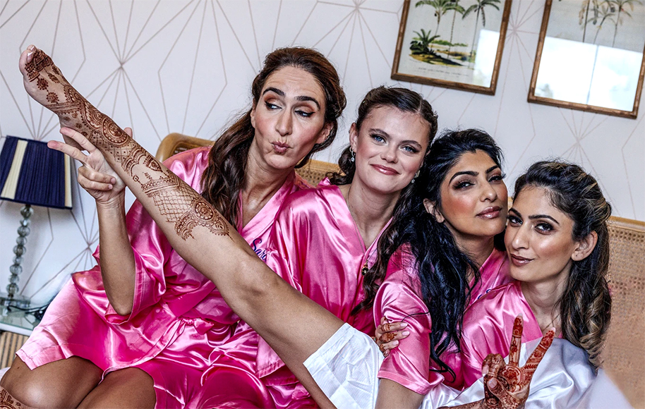 Four women in pink robes sit closely together on a bed, smiling and posing. One woman lifts her leg, showing intricate henna designs on her leg and hand.