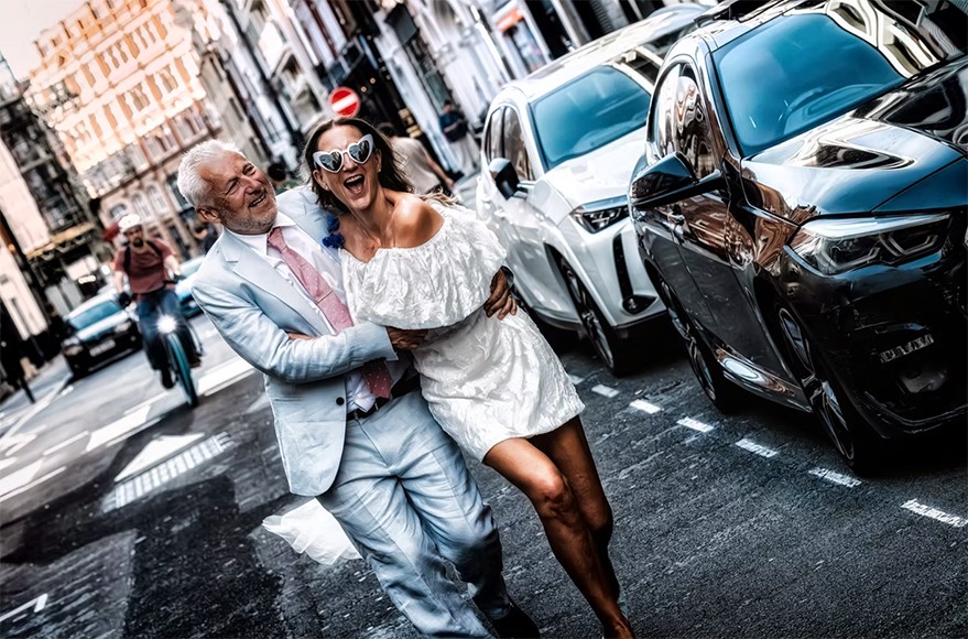 An older man in a light suit and a woman in a white dress with heart-shaped sunglasses laugh and walk together on a city street, with parked cars and a bicyclist in the background.