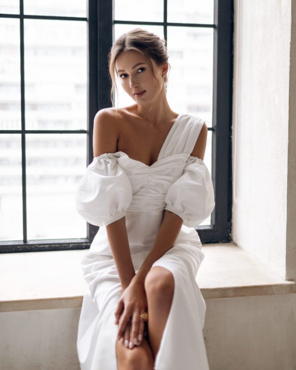 A woman in an off-the-shoulder white dress sitting on a window ledge.