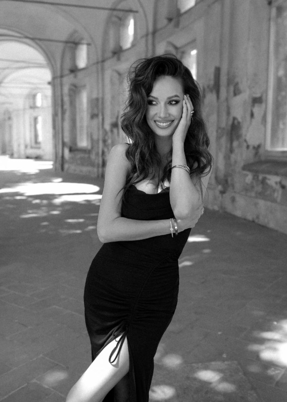 A smiling woman with wavy hair wearing a black dress with a side slit stands in a stone corridor.