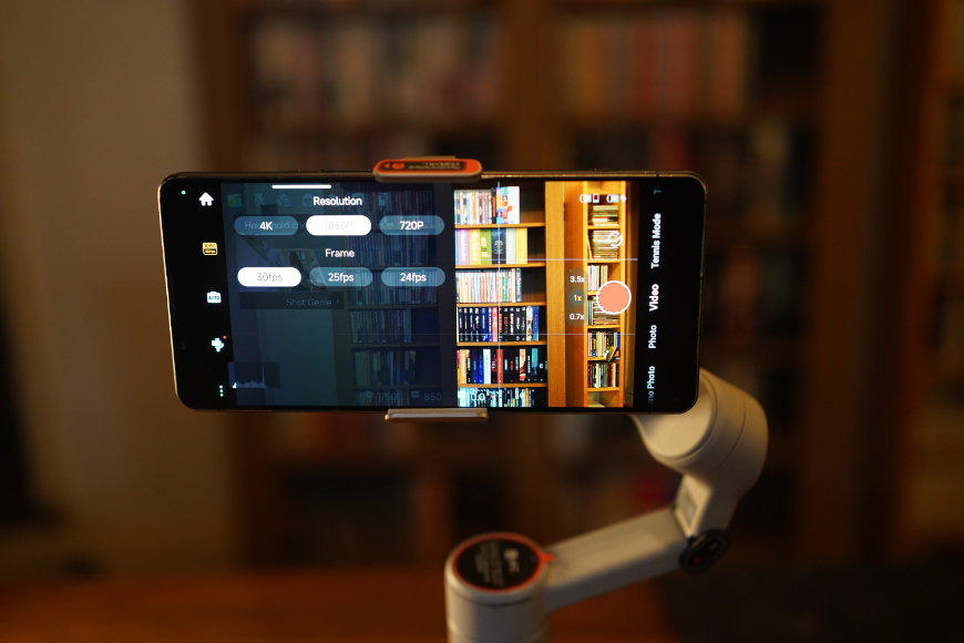 A smartphone mounted on a gimbal, displaying camera resolution and frame rate settings over a live view of a bookshelf.