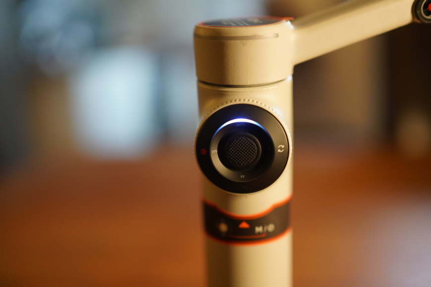 Close-up of a handheld camera gimbal featuring a central joystick and an illuminated status indicator light.