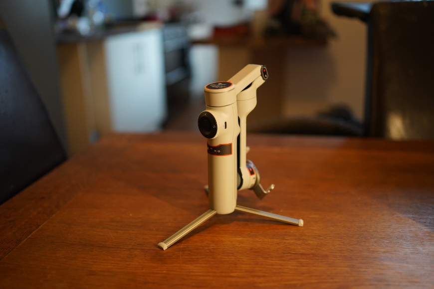 A white handheld gimbal stabilizer standing on a small tripod base on a wooden table.