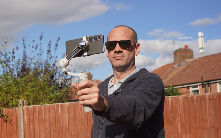A man wearing sunglasses holds a smartphone mounted on a motorized gimbal stabilizer.