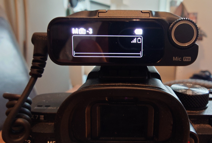 A wireless microphone receiver mounted on a camera, displaying a digital menu with signal and battery levels.