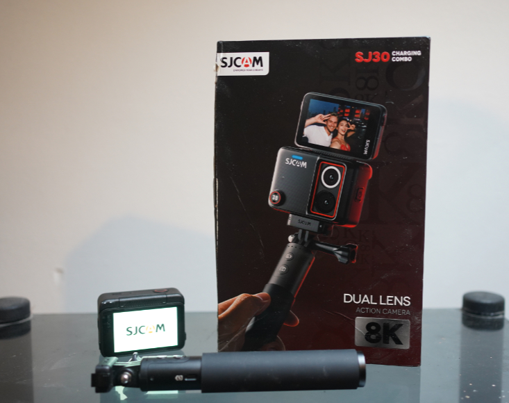 An SJCAM SJ30 action camera and selfie stick displayed in front of its product packaging.