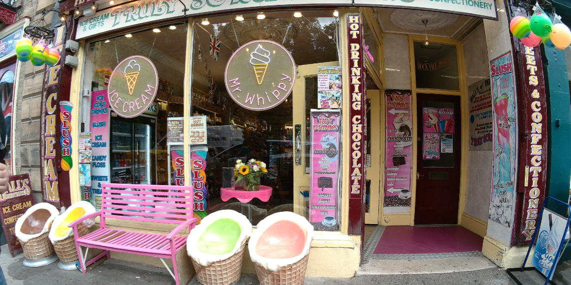 The storefront of an ice cream shop with two giant ice cream cone seats, a pink bench, and posters advertising slushies and hot drinks.
