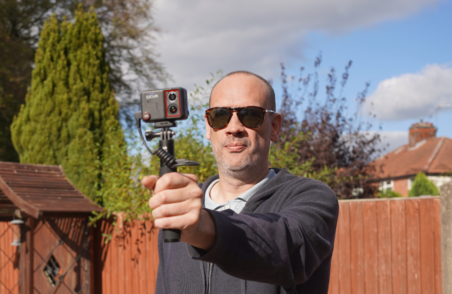 A man wearing sunglasses holds a small action camera on a handheld tripod in front of him.