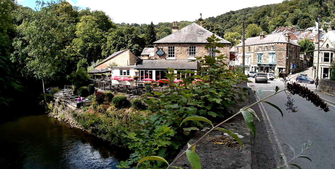 The Fisherman’s Inn pub sits beside a river and a road, surrounded by trees and stone buildings in a valley.