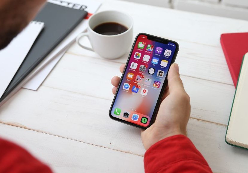 A man holding an iphone x on a desk with a cup of coffee.