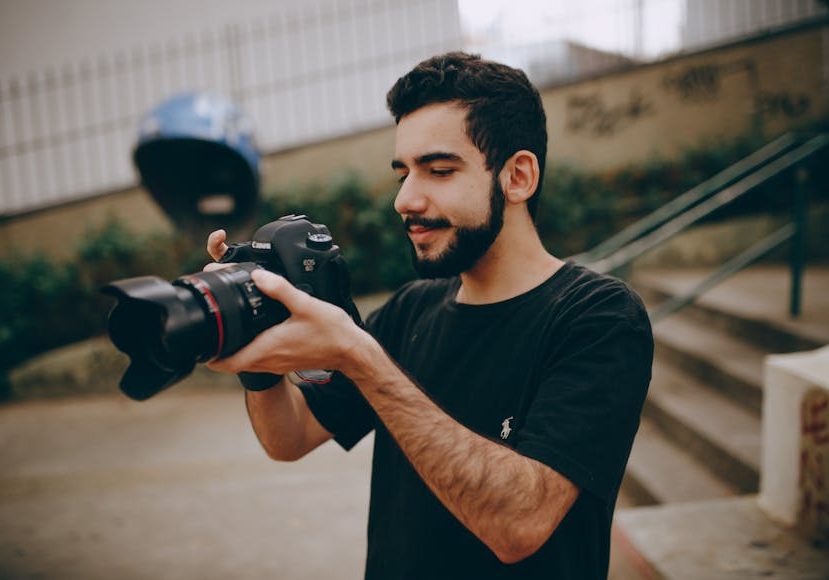 A young man with a beard examining photos on a digital camera outdoors, wearing a black t-shirt, with a blurred background.