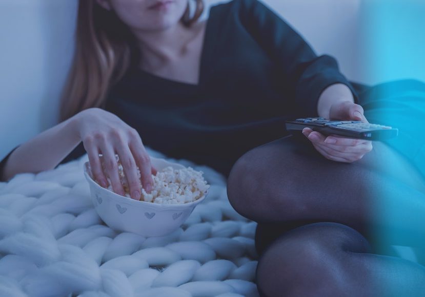 A woman sitting on a bed with a bowl of popcorn and a remote control.