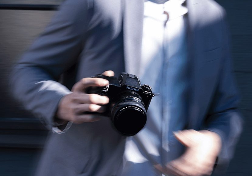a man in a suit and tie holding a Fujifilm gfx100s camera with lens attached.