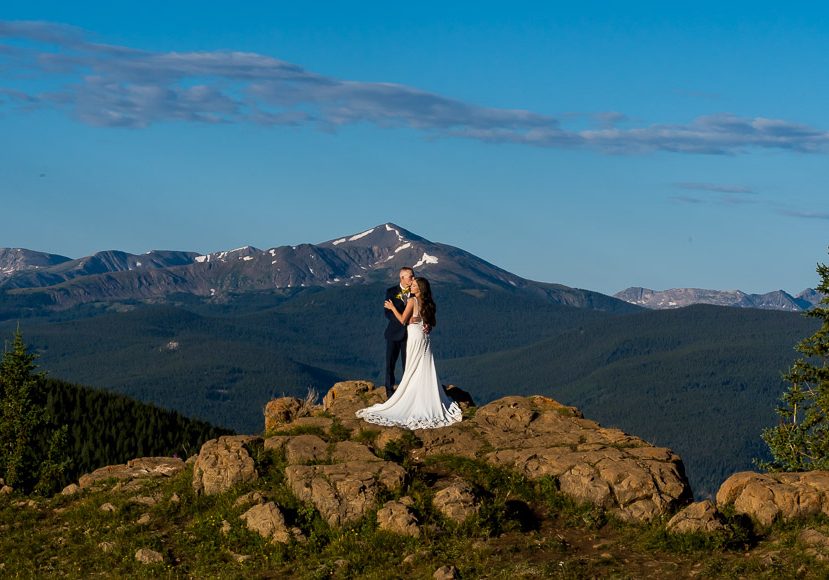 A bride and groom standing on top of a rock with mountains in the background.