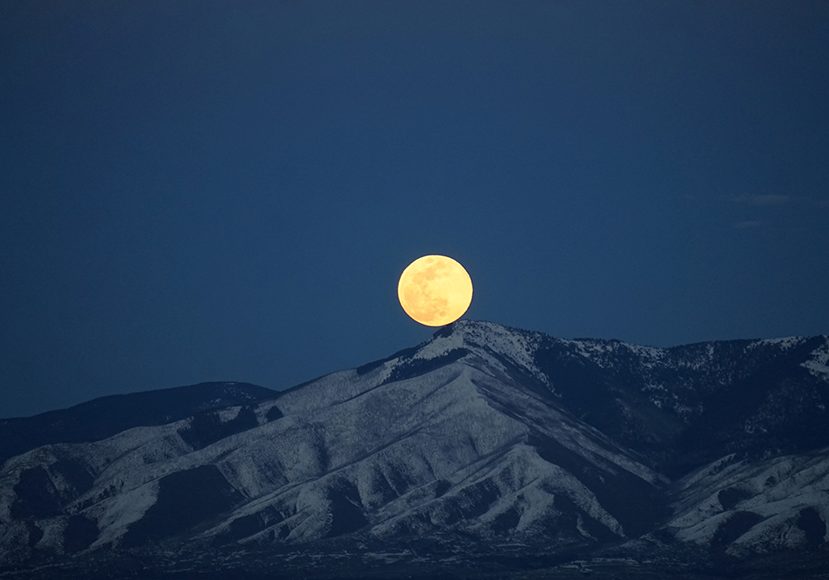 A full moon rises over a snowy mountain range.