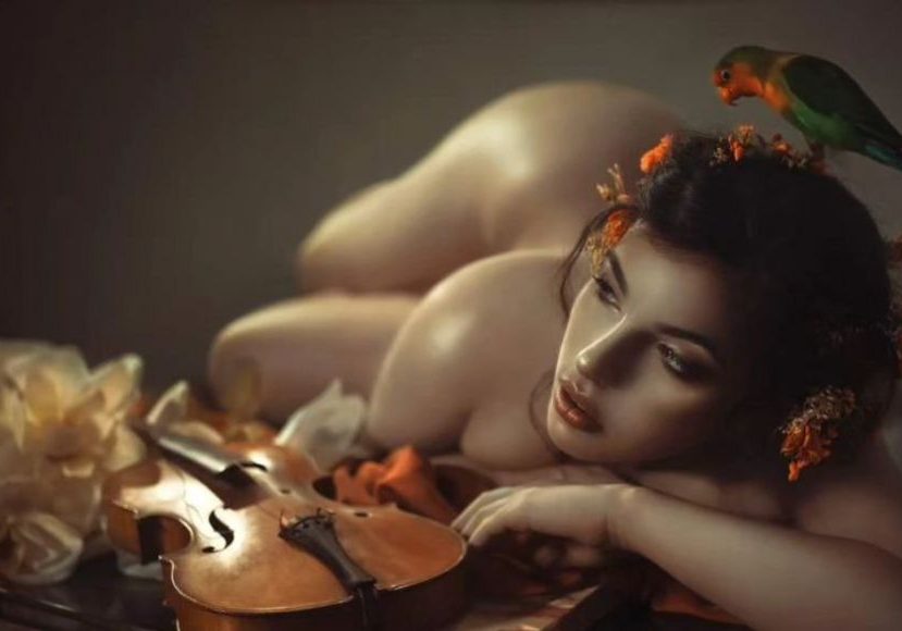 A nude woman laying on a violin with a parrot on her head.