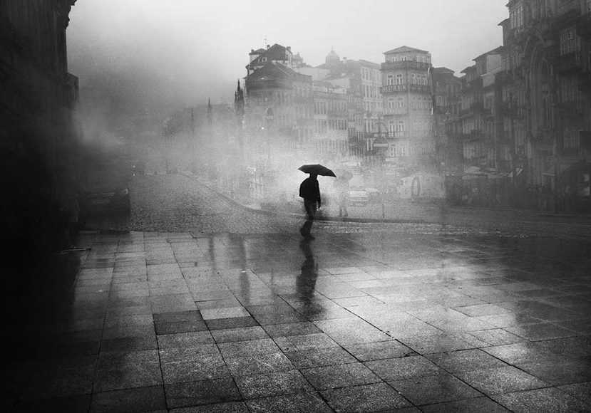 A black and white photo of a person walking down a street with an umbrella.