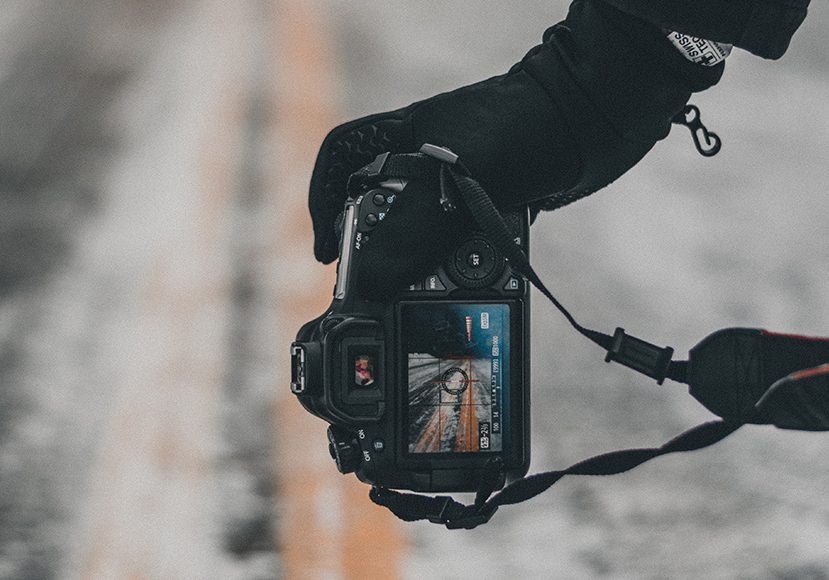 A person holding a camera on a snowy road wearing gloves.