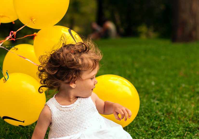 A little girl in a white dress sitting in the grass with yellow balloons.