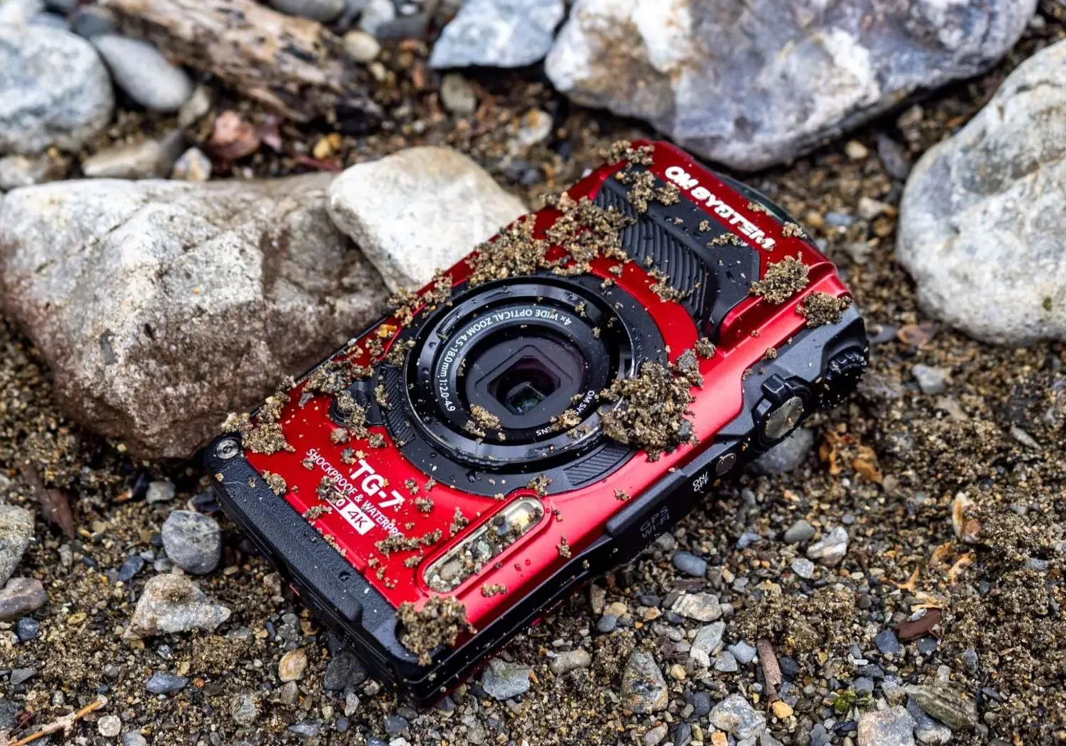 An OM System TG-7 camera is laying on a rock in the mud.