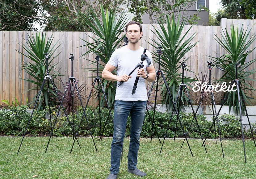 A man stands in a garden holding a camera tripod, surrounded by various tripods of different sizes, with plants in the background.
