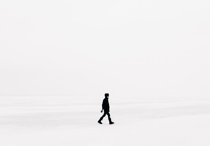 a black and white image of a man walking across a snowy field.